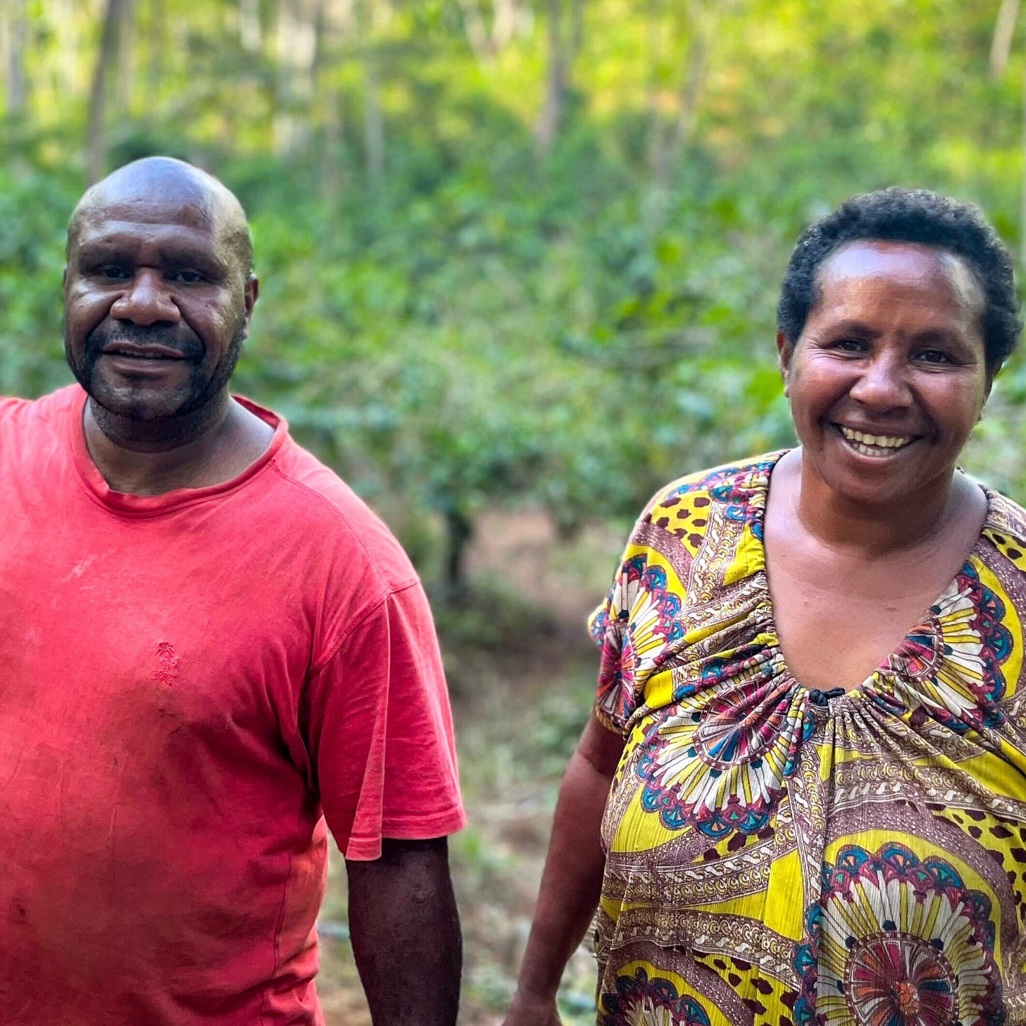 Two people standing in a forested area, one wearing a red shirt and the other in a patterned outfit.